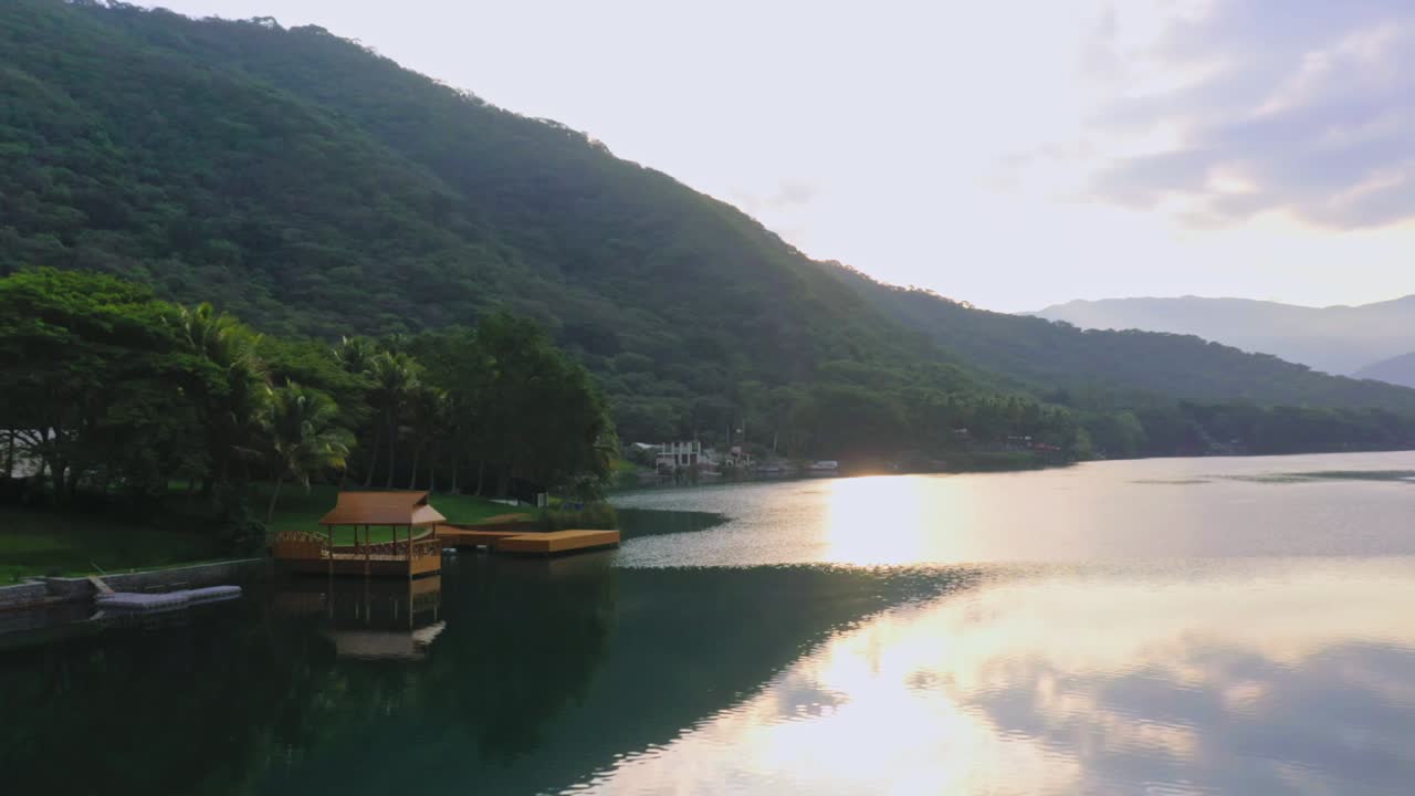 Tranquil scene of a forested lakeshore at sunrise. Calm waters reflect the golden sky, mountains in the backdrop enhancing serenity. Perfect for nature, travel, and relaxation themes