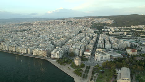 cinematic-wide-drone-aerial-shot-of-northern-Greece-city-of-Thessaloniki-with-the-sea-and-city-and-white-tower-in-view-at-sunset,-moving-backwards-in-4k