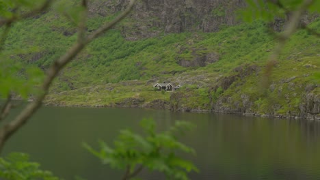 Small-cottages-by-the-serene-Agavatnet-Lake-in-lush-Lofoten,-Norway,-captured-through-trees