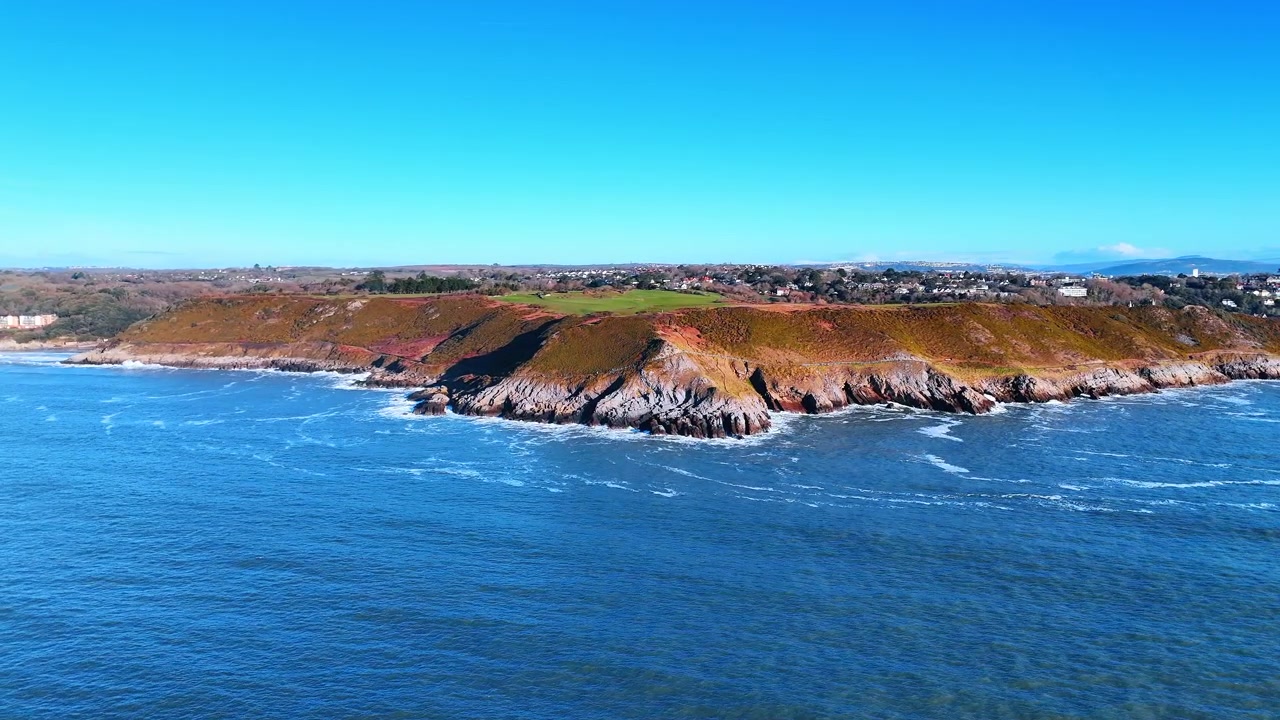 Vista Aérea de Promontório Costeiro com Ilhota de Farol Distante. Um promontório arborizado estende-se em direção a pequenas ilhotas rochosas com um farol, enquanto a baía se abre para uma costa distante enevoada.