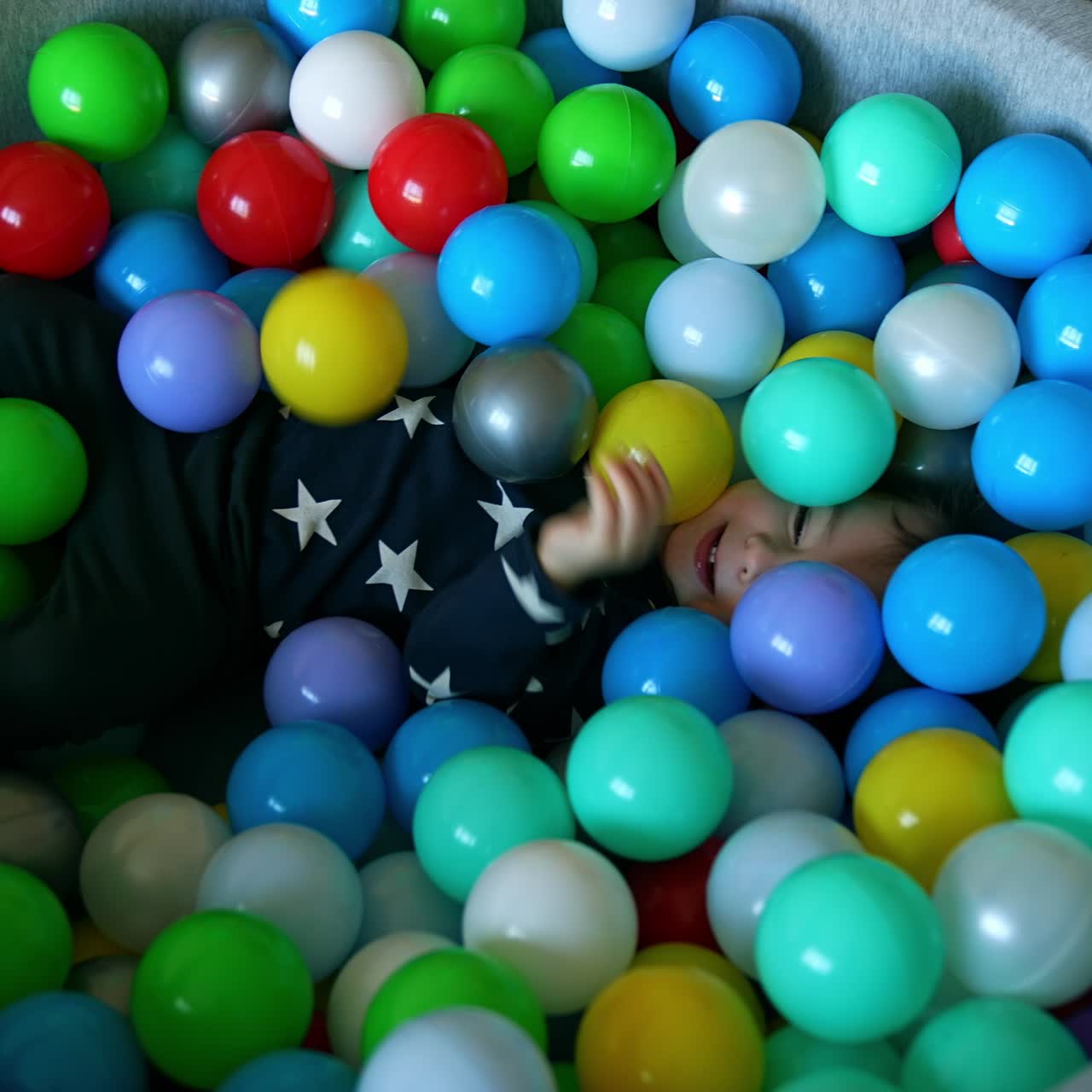 Adorable active toddler jumps into the dry basin with balls. Happy funny kid deepens into the toys. Top view