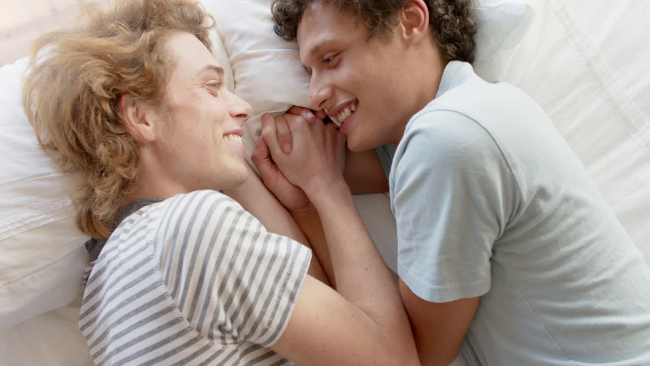 Felice coppia di uomini gay di diversa etnia sdraiata sul letto che si tiene per mano a casa, al rallentatore