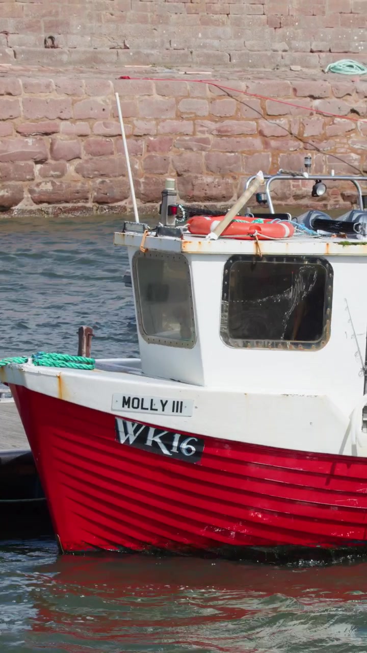 Una piccola barca da pesca rossa si muove lentamente vicino a una banchina in pietra in pieno giorno, acque tranquille