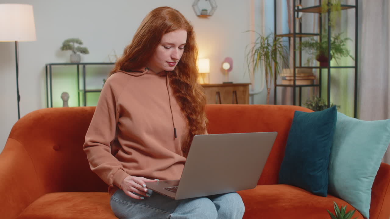 Happy young woman lady sitting on sofa opens laptop and working on project in living room at home