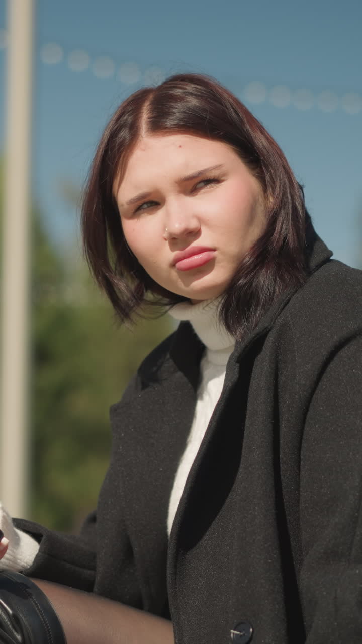 흑색 코트와 색 스웨터를 입고 밖에서 앉아있는 사려 깊은 여성, 그녀의 반지 장식 손이 보이고, 배경에는 흐릿한 초록색과 나무가 있습니다.