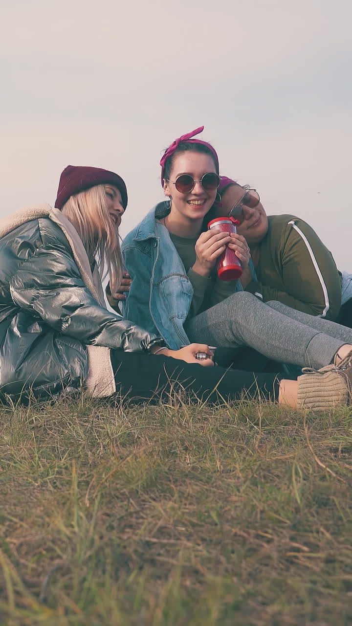 cheerful young women cover friend with denim jacket sitting on grass against clear sky in autumn evening low angle shot