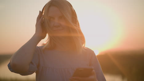 tranquil scene of a woman in a blue gown, enjoying a sunset with a black headset, gently holding it while using her smartphone, under a glowing sun effect