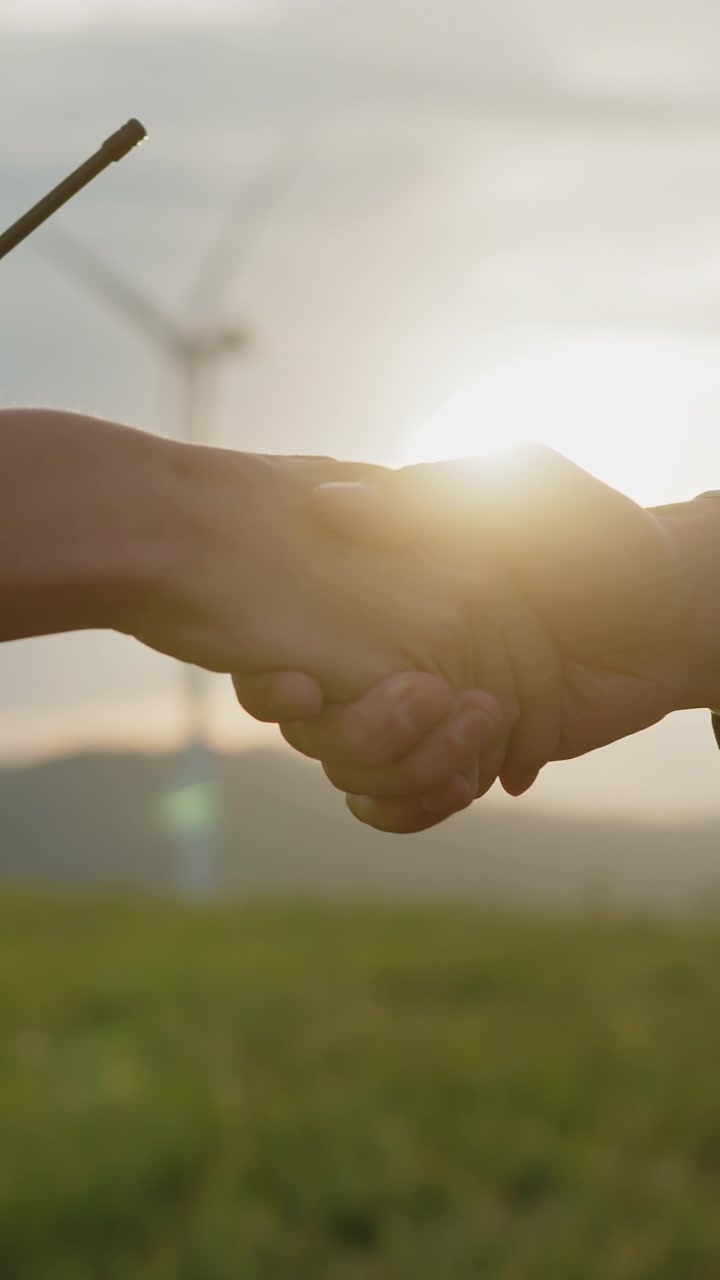 Two hands shake in front of wind turbines at sunset symbolizing renewable energy partnership and