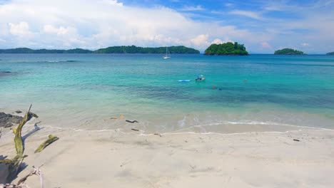 Low-Strafe-Across-White-Sand-Beach-Edge-with-Sailboat-and-Swimmers