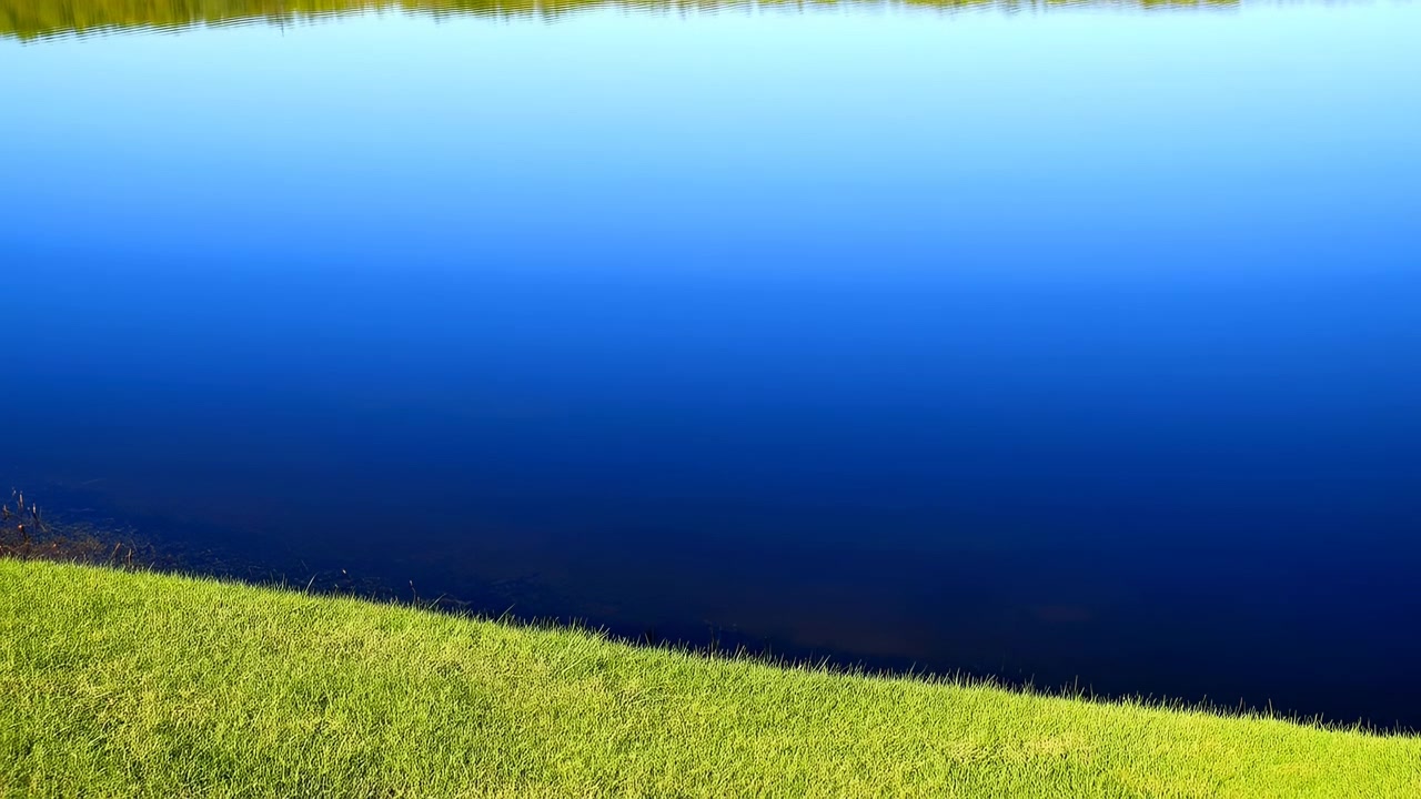 Oever van blauw meer met levendige grashelling, glashelder diepblauw water en verre boomgrens, zachte bries, zacht