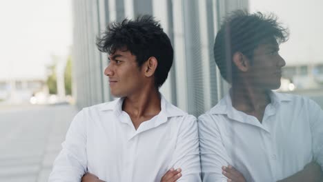 indian young guy crossing his arms, leaning against a glass wall on the street