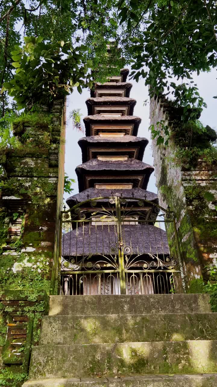 Lodret billede af en traditionel balinesisk tempelpavillon med udskårne stendetaljer, ceremonielt alter og stråtag set gennem en klassisk todelt port inde i en fredfyldt gårdhave på Bali, Indonesien