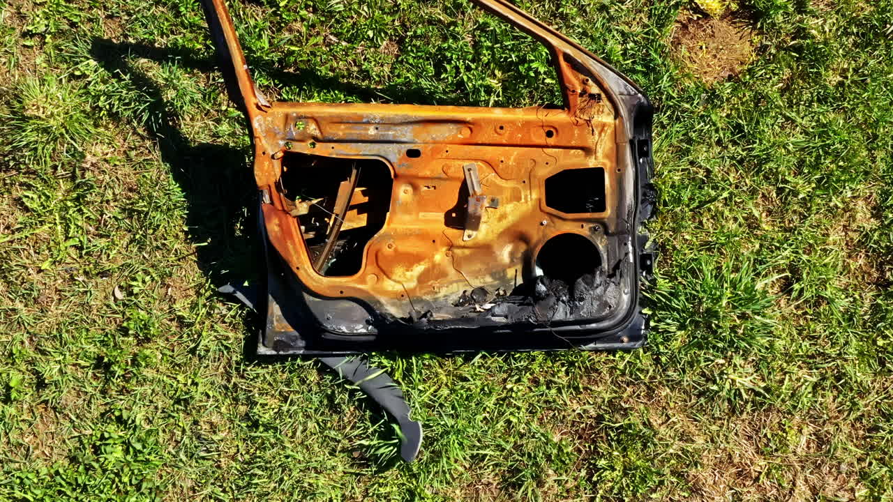 Burned rusted car door panel discarded in grassy field under daylight