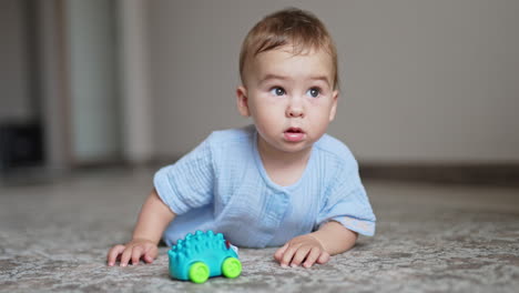 lovely baby boy in blue clothes lies on his belly on the floor. cute child has a toy in front of him but looks around with interest.