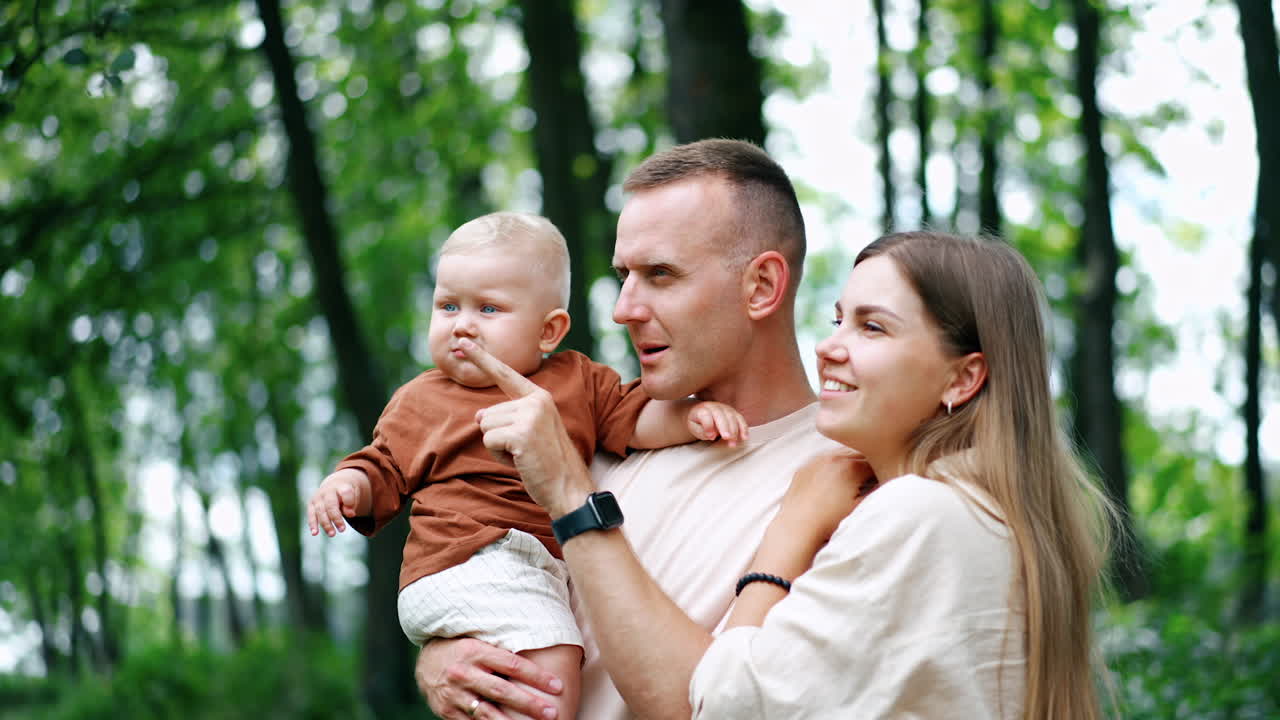 Caucasian parents stand outdoors with their toddler baby boy. Dad talks to his son and points at something and kid repeats the move.