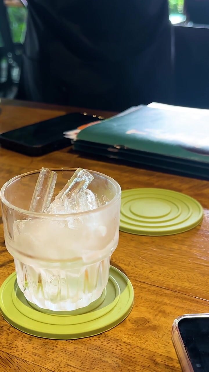 A hand places a glass filled with ice on a wooden table in a restaurant setting, under natural lighting