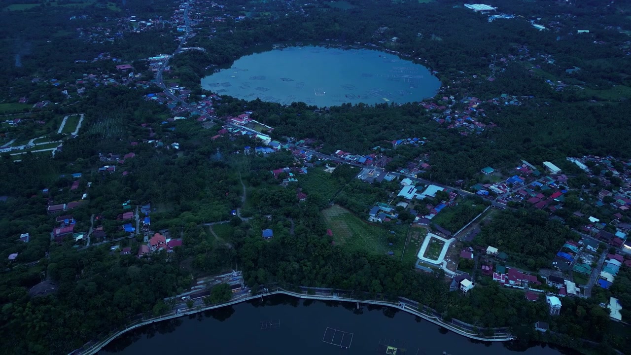 Alacakaranlıkta Filipinler'in Laguna eyaletindeki San Pablo Şehri'nde Sampaloc Gölü, Bunot Gölü, yemyeşil tropikal bitki örtüsü, çevreleyen şehir manzaraları ve San Cristobal Dağı'nı sergileyen geri çekilen drone görüntüsü.