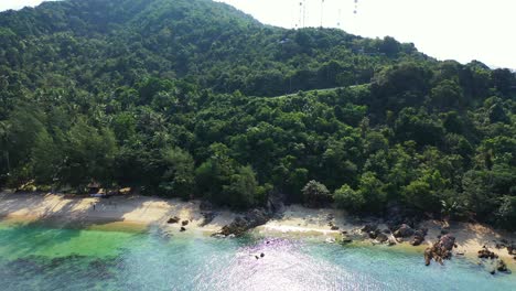 Green-hill-with-lush-vegetation,-antennas-on-the-top-and-rocky-coastline-washed-by-calm-clear-water-reflecting-sunshine-in-Thailand