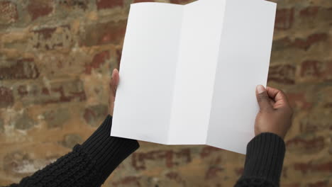 video of hand of african american man holding white blank page, copy space over brick wall