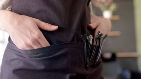 Midsection-of-caucasian-female-hairdresser-in-apron-with-hands-and-hair-clips-in-pocket,-slow-motion