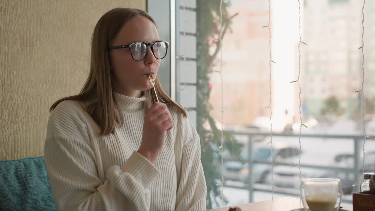 close up of young woman in white sweater slicing chocolate cake then licking spoon happily at cafe table near window with snowy street and decorative string lights