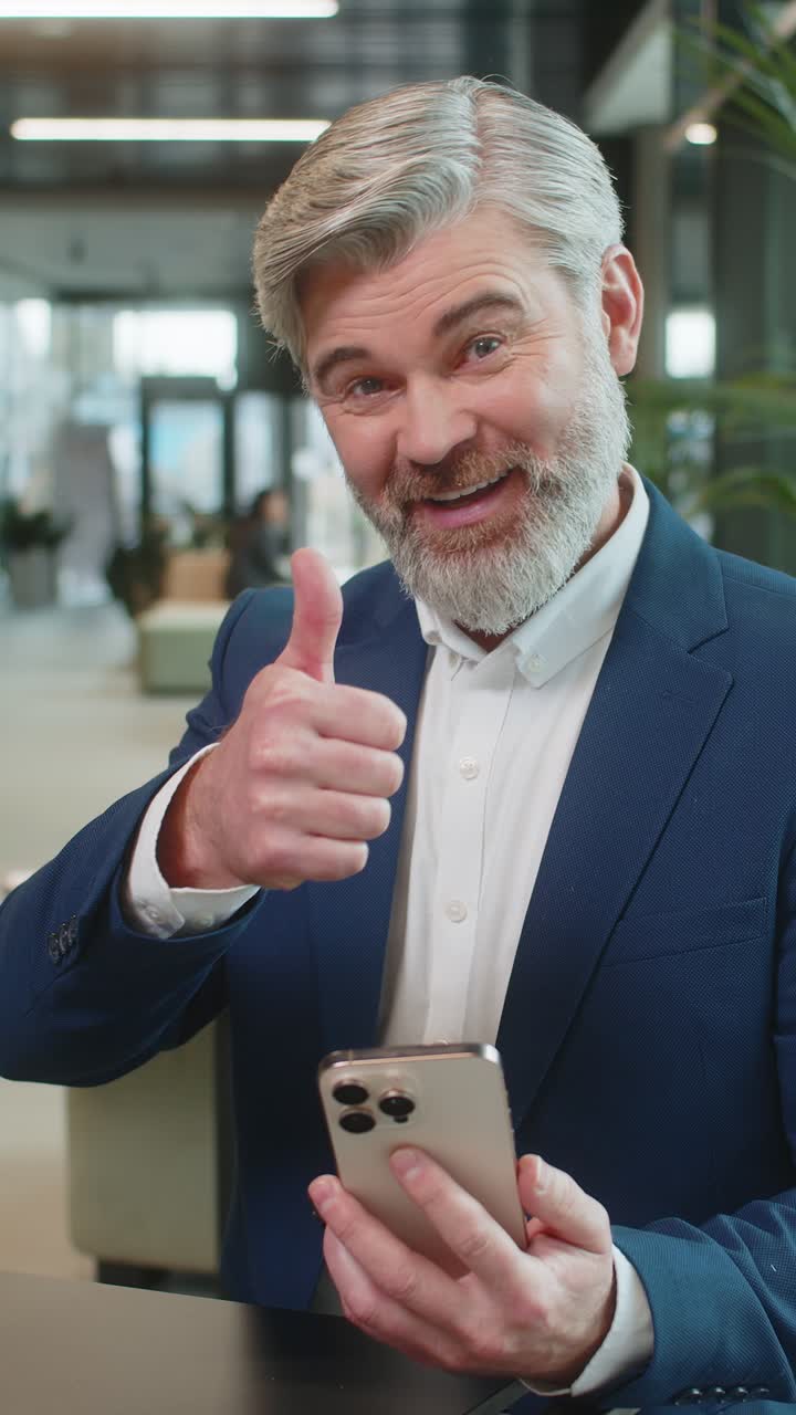Smiling mature businessman in suit recommends using the phone app sitting in modern office lobby