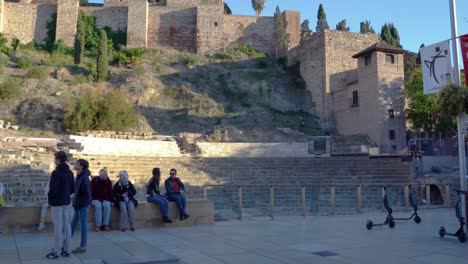 people walk in front of teatro romano de malaga, alcazaba in background, spain, costa del sol