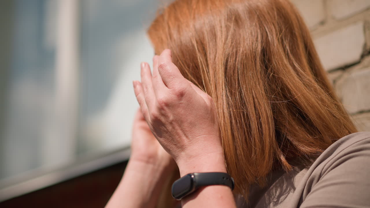 Tense woman with long brown hair lean against sunlit brick wall, eyes lowered in discomfort, preparing to wear sunglasses, sunlight highlighting expression of fatigue