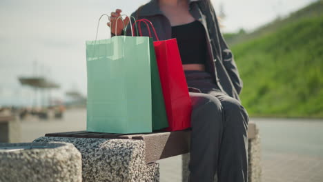 lady in grey clothing drops green, mint, red shopping bags on bench while sitting, with a slight view of greenery hill in the background, and distance blur object