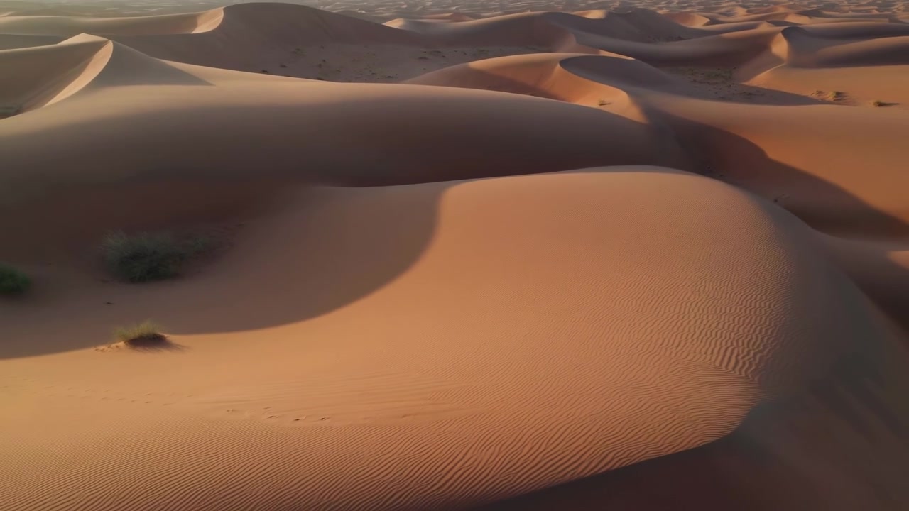 Dune del deserto a strati con sabbia fine increspata, calde tonalità dorate, ampie fasce d'ombra e pendii ondulati in un tranquillo paesaggio arido, viste da un punto di osservazione obliquo elevato sotto una luce soffusa e bassa.