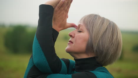 close-up side view of middle-aged woman with hand twist, touching her forehead while eyes are closed, practicing yoga outdoors in a misty grassy field, with blurred trees in the background