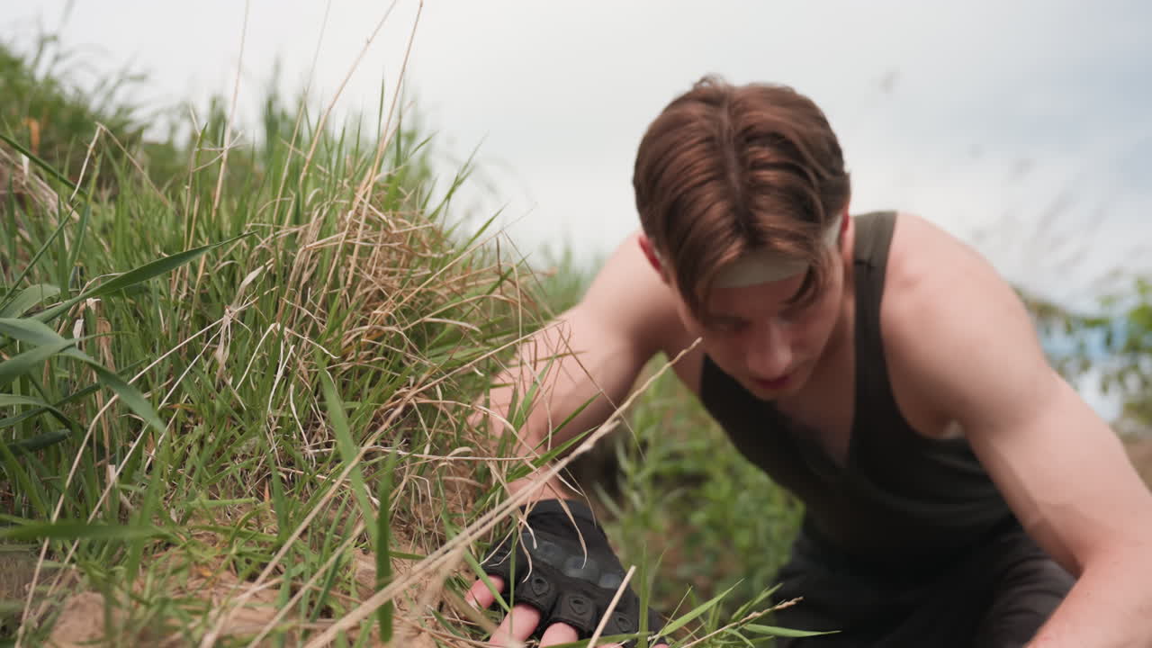 warrior steadying hand on grassy sand mound while sliding boot onto foot for secure grip before navigating steep river bank during remote wilderness trek under pale cloudy sky with calming atmosphere