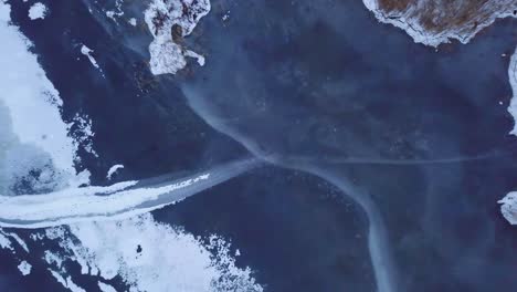 Aerial-birdseye-view-of-frozen-lake-Liepaja-during-the-winter,-blue-ice-with-cracks,-dry-yellowed-reed-islands,-overcast-winter-day,-ascending-drone-shot