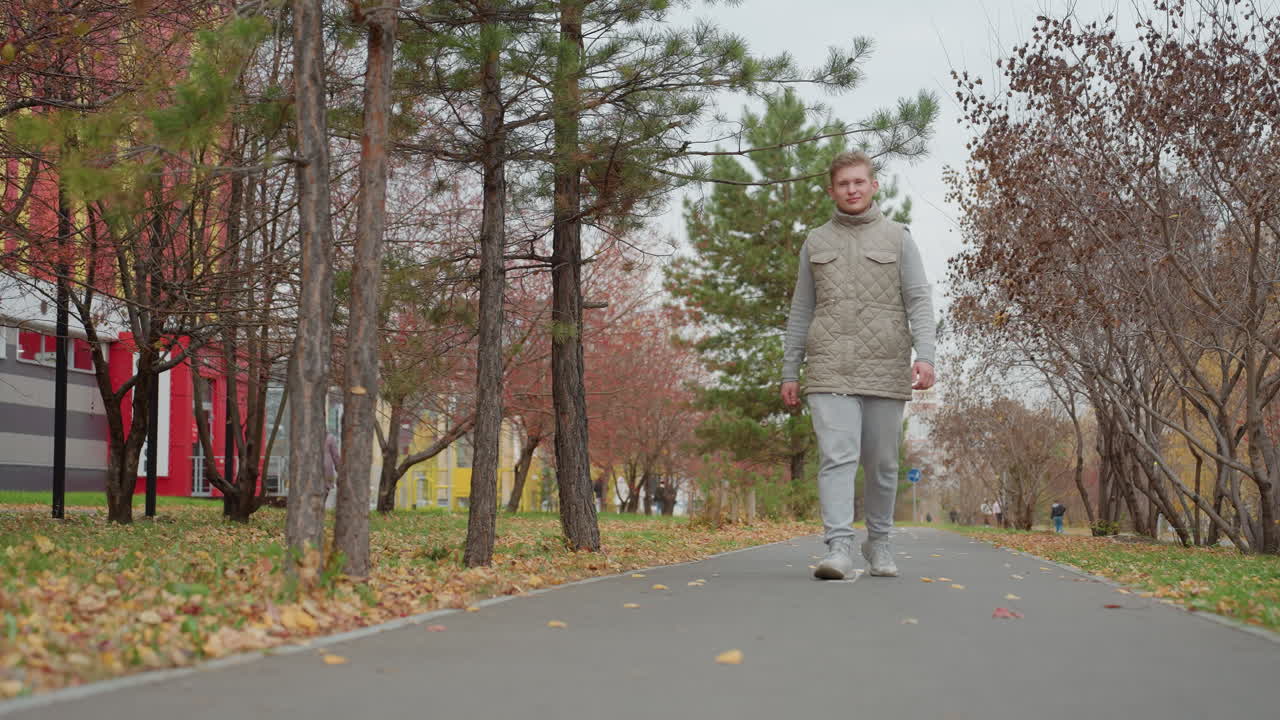 Business man in jacket walks confidently along tarred path as cool breeze moves through autumn park with dry leaves scattered around while another person walks opposite side in urban background