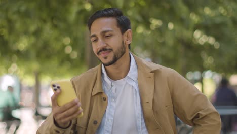 portrait of smiling muslim man sitting at outdoor table on city street looking at mobile phone