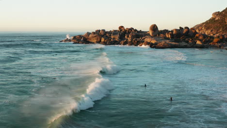 aerial flying along with the waves with surfers in the background in llandudno, cape town, south africa