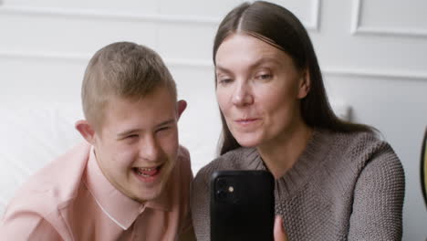 close-up view of a boy with down syndrome and his mother watching something on smartphone lying on the bed in the bedroom at home