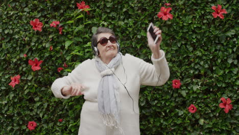 Porträt-Einer-älteren-Kaukasischen-Frau,-Die-Tanzt-Und-Verspielt-Tanzt,-Mit-Erhobenen-Armen-Und-Musik-über-Kopfhörer-Mit-Sonnenbrille-Im-Gartenhintergrund-Im-Freien-Hört