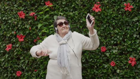 Retrato-De-Una-Anciana-Caucásica-Bailando-Disfrutando-De-Un-Baile-Juguetón-Con-Los-Brazos-Levantados-Escuchando-Música-Con-Auriculares-Usando-Gafas-De-Sol-En-El-Fondo-Del-Jardín-Al-Aire-Libre