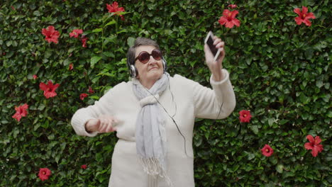 retrato de una anciana mujer caucásica bailando disfrutando de la danza lúdica brazos levantados escuchando música en auriculares usando gafas de sol al aire libre jardín de fondo