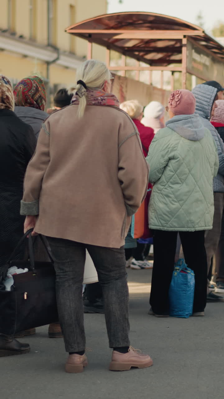 elderly caucasian women waiting at market group of seniors with headscarves and coats queue along pavement near vendor stalls carrying bags and trolleys muted autumn light candid backview communal