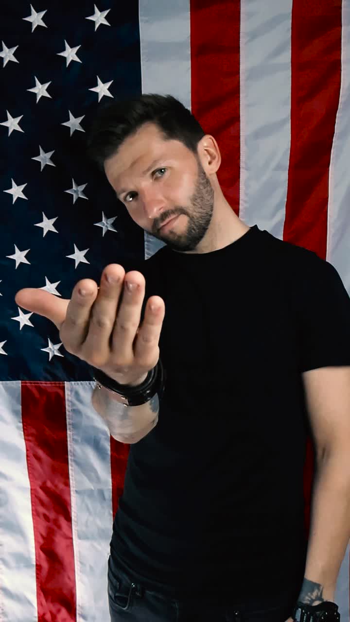 Tattooed man pointing to the camera with his index finger in slow motion and does come on or come at me wave with his hand, invites. Standing in front of a vertical American flag that is moving