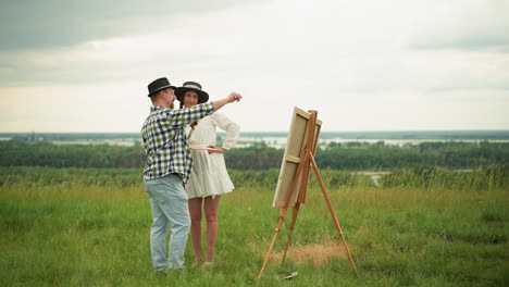 a man in a black hat and plaid shirt stands close to a woman in a white dress in a grassy field. they are engaged in a discussion or observation, with a drawing board set up in front of them