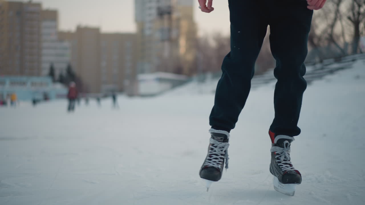 Close up of people skating on frozen lake near urban buildings, ice surface with skate marks and snow, focus on legs wearing skates, blurred background featuring skaters and cityscape