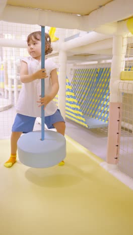 a determined 5-year-old girl falls off a soft light blue tire swing on a bright indoor playground every time, but tries to hold on, showing resilience despite the difficulties