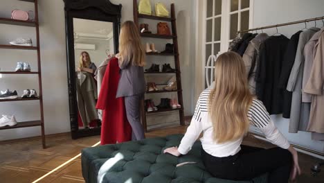 Long-Haired-Young-Blonde-Female-Is-Trying-A-New-Grey-And-Red-Coats-While-Standing-In-Front-The-Mirror