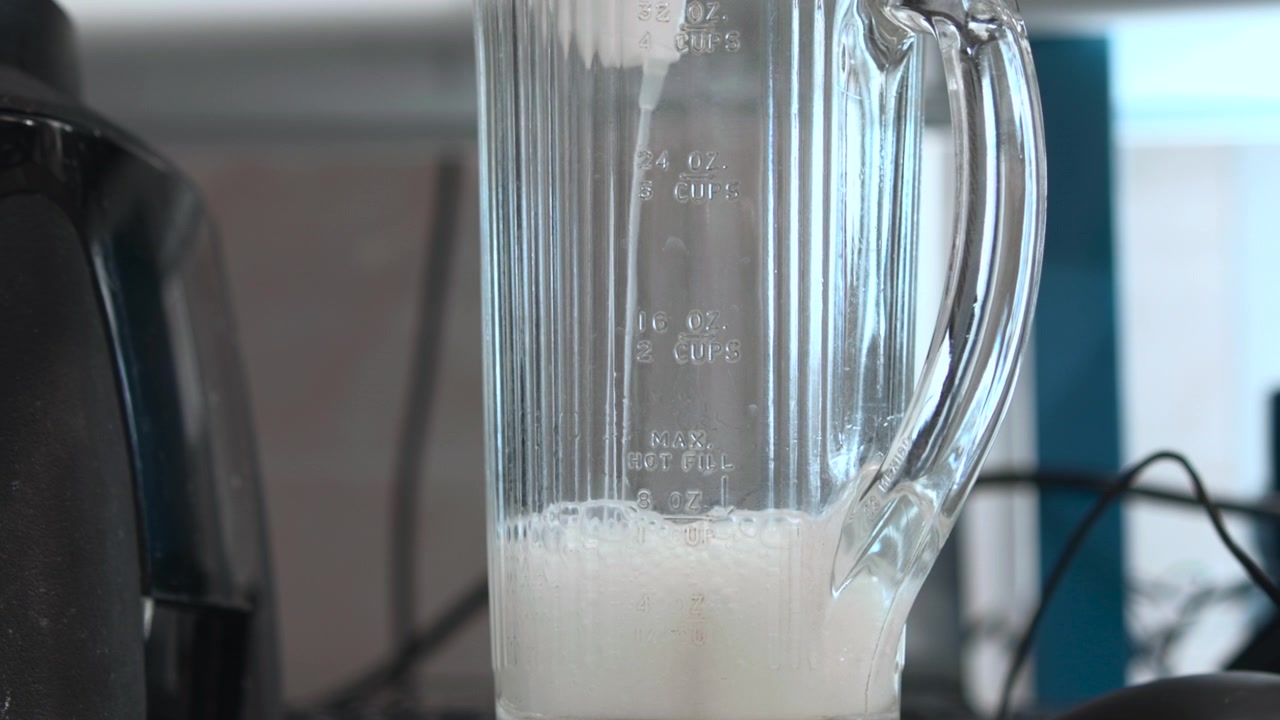 Primo piano all'altezza degli occhi di una caraffa di vetro trasparente di frullatore parzialmente riempita con liquido bianco spumoso, bolle che salgono e si depositano accanto a marcature di misurazione in rilievo, lati a coste e una luce diurna morbida e fredda