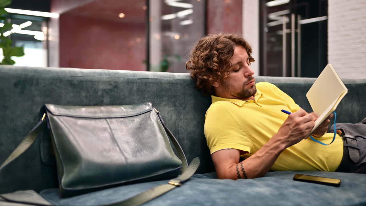 Man in yellow shirt lying on a couch and writing in a notebook