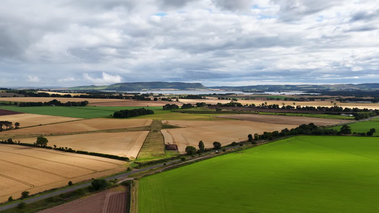 Drone camera glides above patchwork farmland with green and golden fields under cloudy summer skies, capturing rural landscape and agricultural patterns in natural light