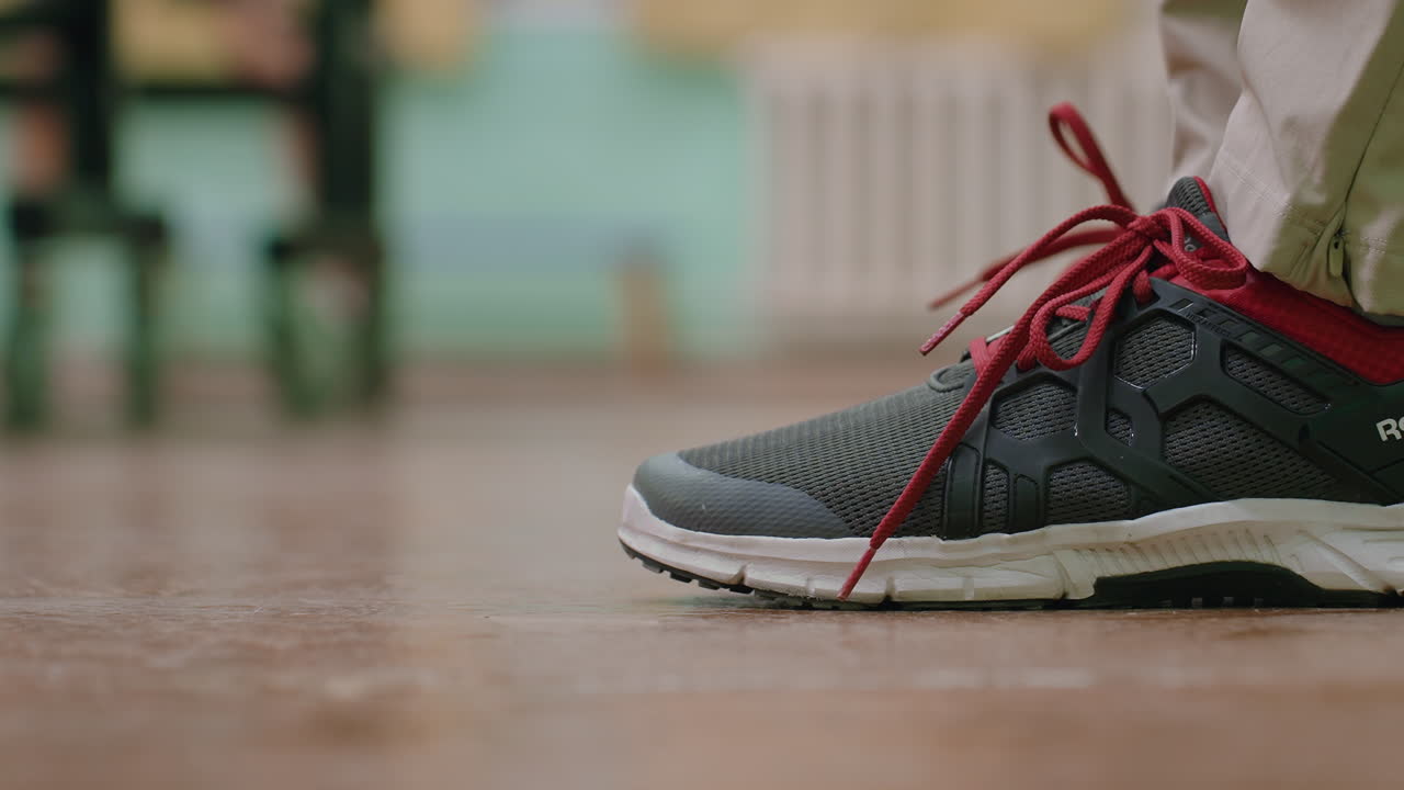 Close up of tennis player wearing dark sneaker with bright red laces standing on indoor court, beige pants slightly visible, moment highlighting detail, and preparation in sports environment